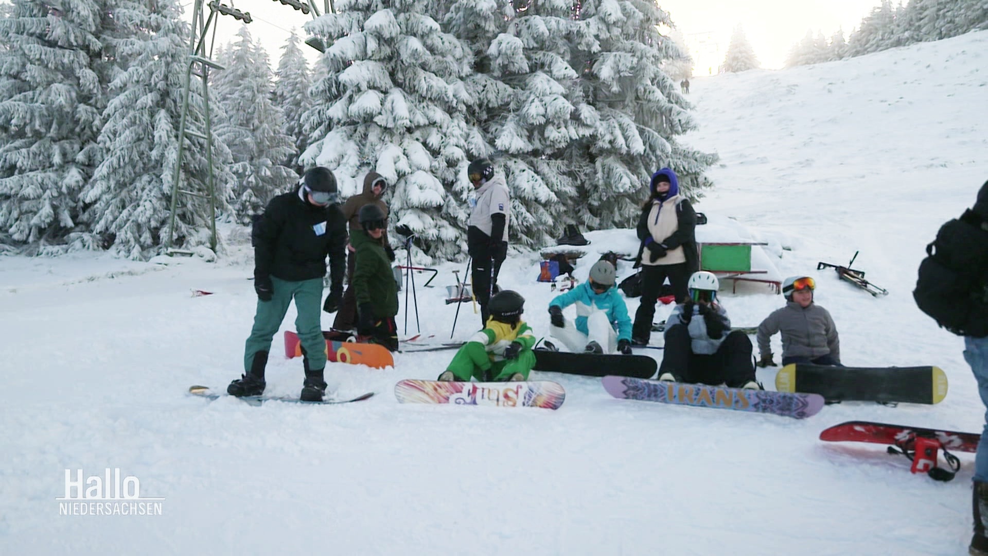 Eine Gruppe Snowboarder und Snowboarderinnen rastet auf der Pist vor einer dick verschneiten Tanne. © Screenshot 