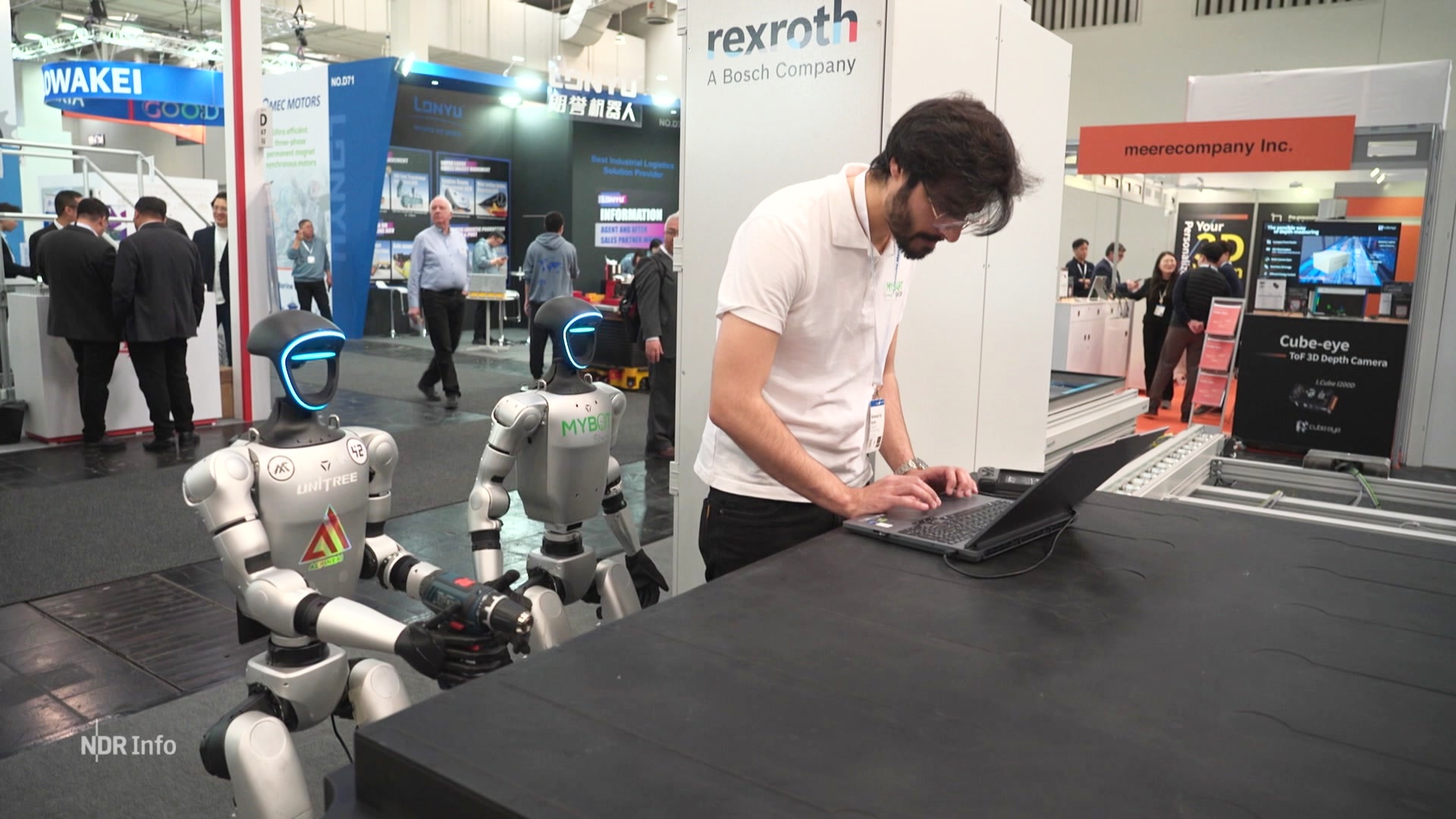 Ein Mann arbeitet an einem Tisch, neben ihm stehen zwei Roboter. © Screenshot 
