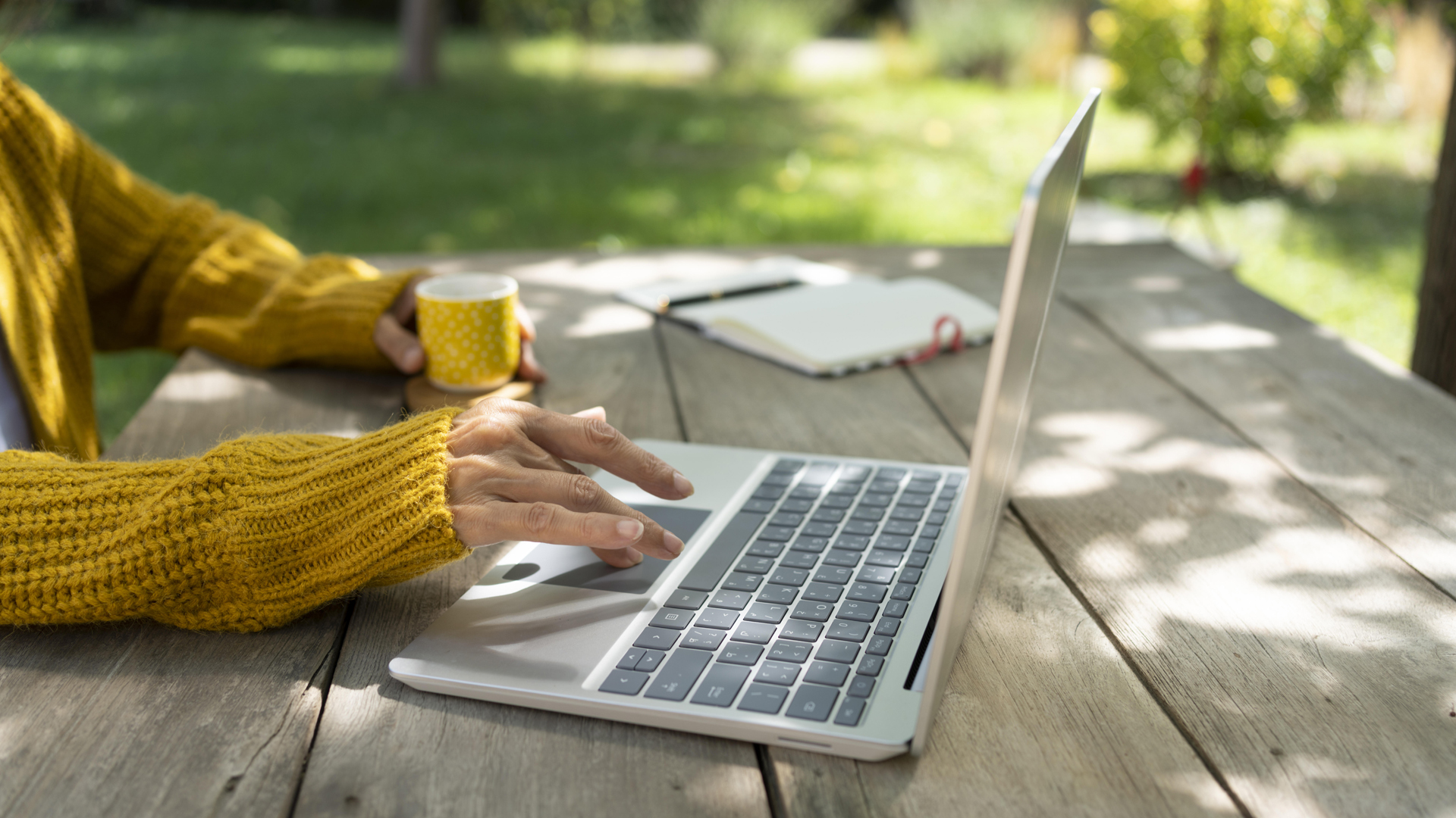 Der Arm einer Frau bedient einen Laptop, der auf einem Tisch in einem Garten steht, während die andere Hand einen Becher hält. © picture alliance / Westend61 | Svetlana Karner 