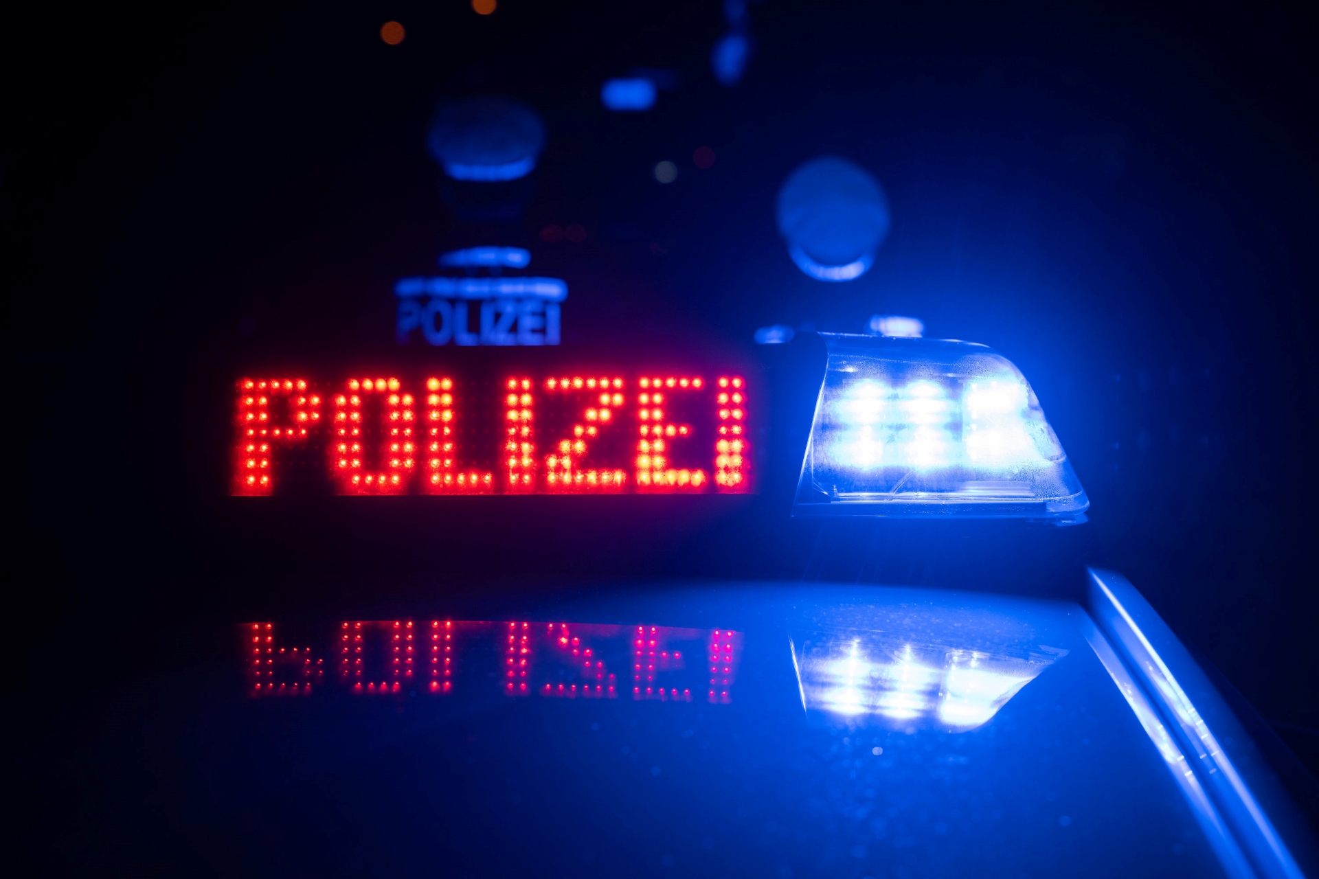 Zwei Polizisten stehen im Dunkeln mit dem Rücken zu ihrem Einsatzfahrzeug, auf dem Dach des Polizeiautos blinkt in roten Buchstaben der Schriftzug "Polizei". (Themenbild) © dpa-Bildfunk Foto: Marijan Murat