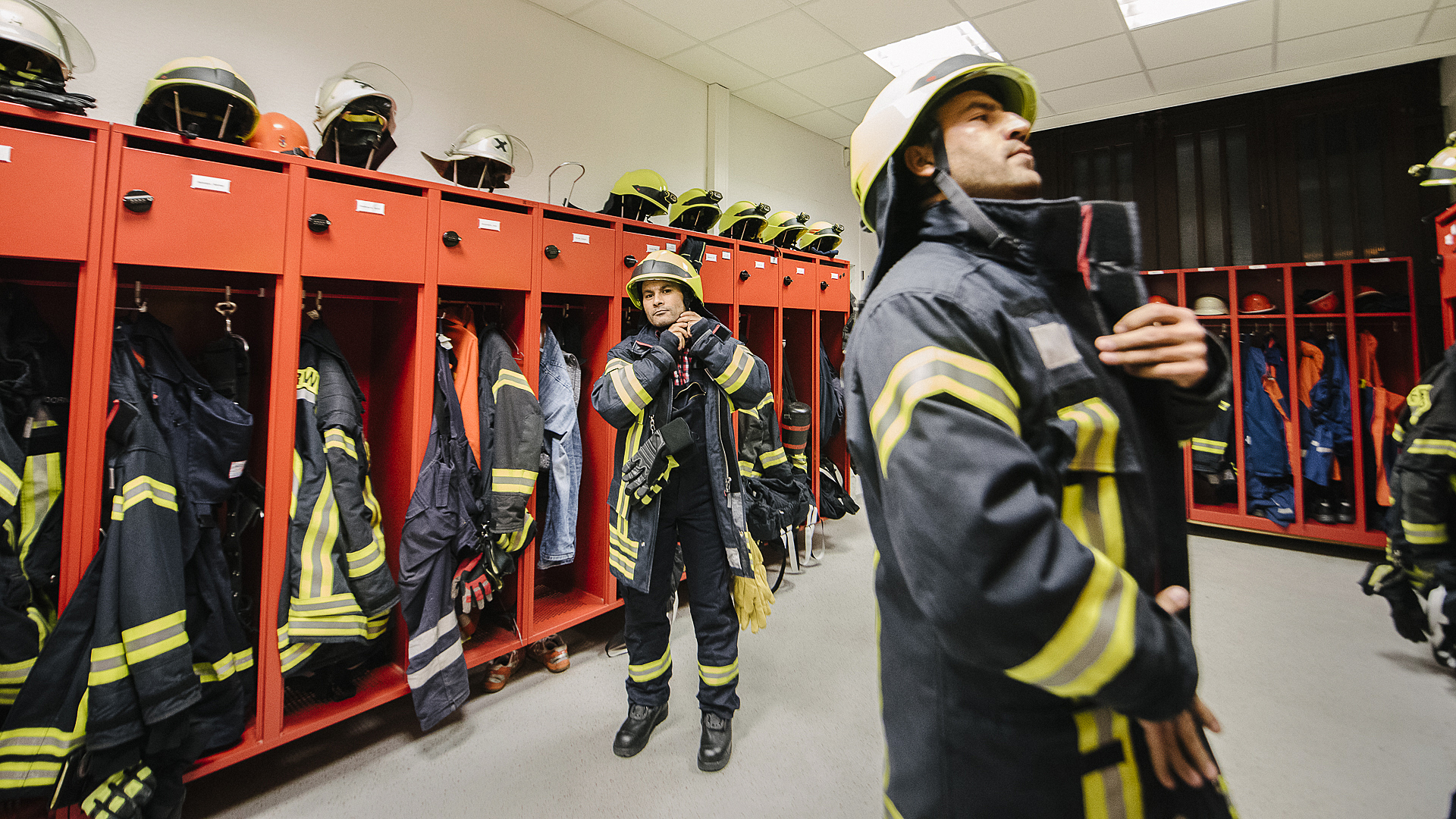 Zwei Feuerwehrmänner ziehen sich in einem Umkleideraum ihre Einsatzkleidung an. © NDR Foto: Julius Matuschik