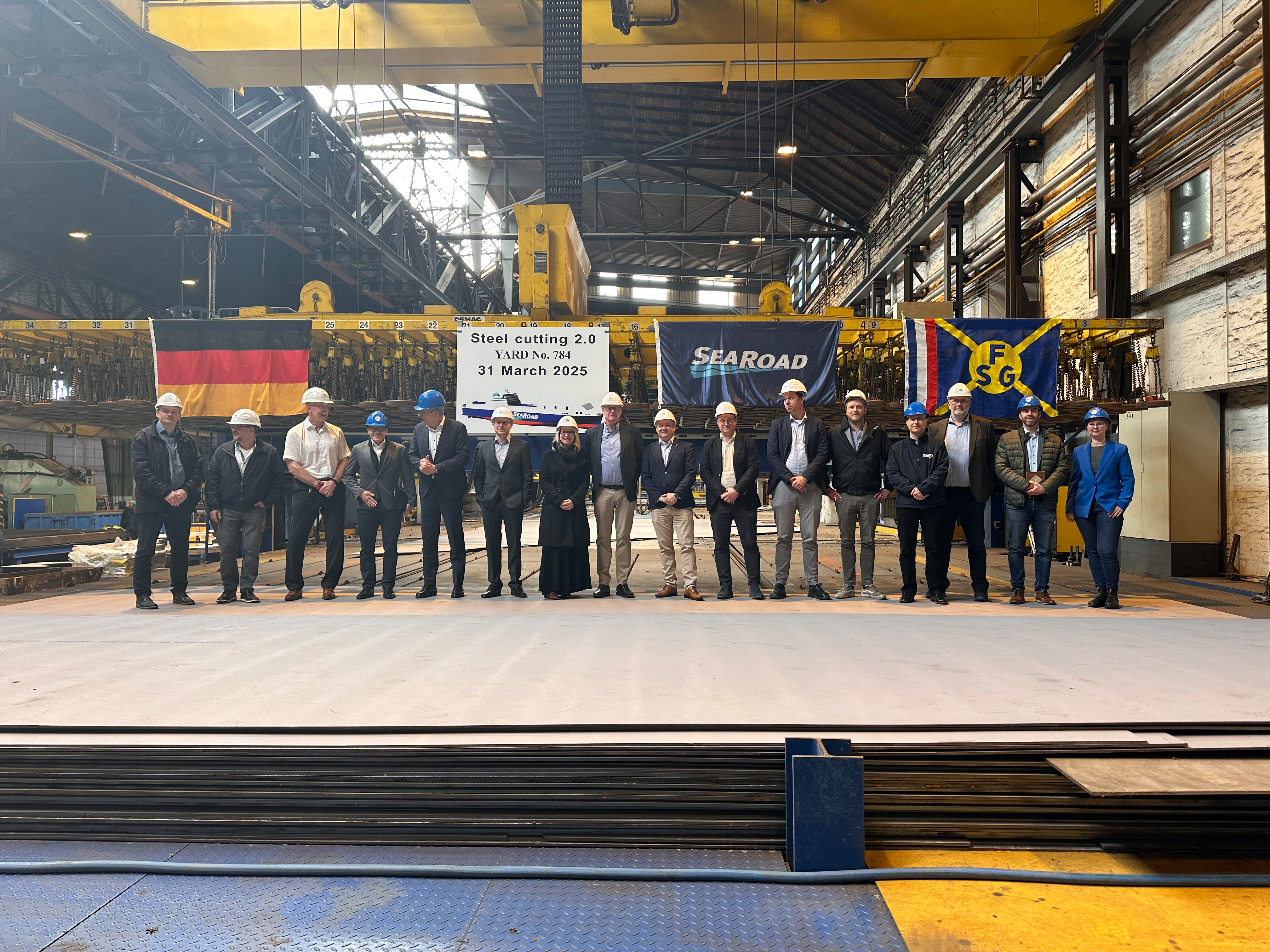 Führungskräfte von FSG und Searoad-Reederei stehen am 31.3.2025 in einer Produktionshalle, in dem ein Plakat auf den Start des Stahlschnitts hinweist. © FSG Shipyard GmbH 
