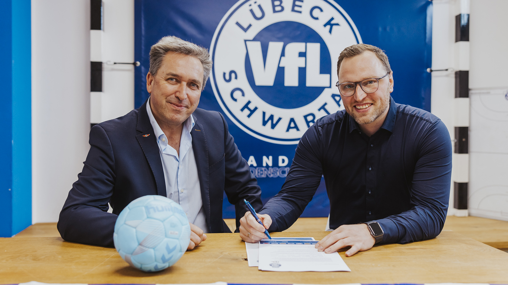 Zwei Männer sitzen an einem Tisch auf dem ein Fußball liegt und lächeln in die Kamera. Zwei Männer sitzen an einem Tisch auf dem ein Fußball liegt und lächeln in die Kamera. © VfL Lübeck-Schwartau